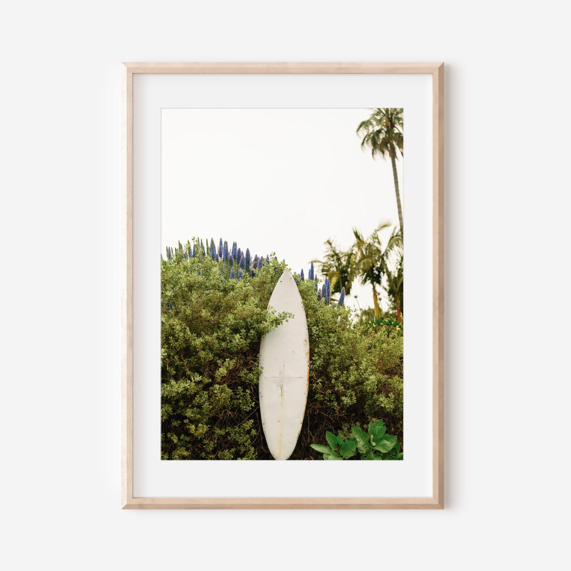 White Surfboard Leaning on a Lush Hedge With Palm Trees Poster